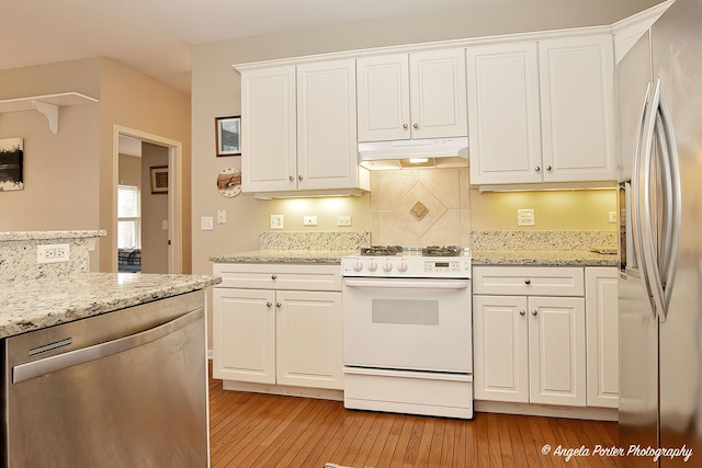 kitchen with under cabinet range hood, stainless steel appliances, white cabinets, light stone countertops, and light wood finished floors