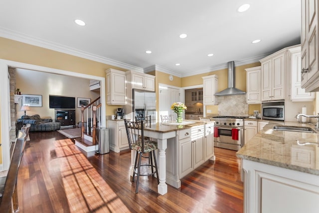 kitchen with a sink, a center island, appliances with stainless steel finishes, wall chimney range hood, and light stone countertops