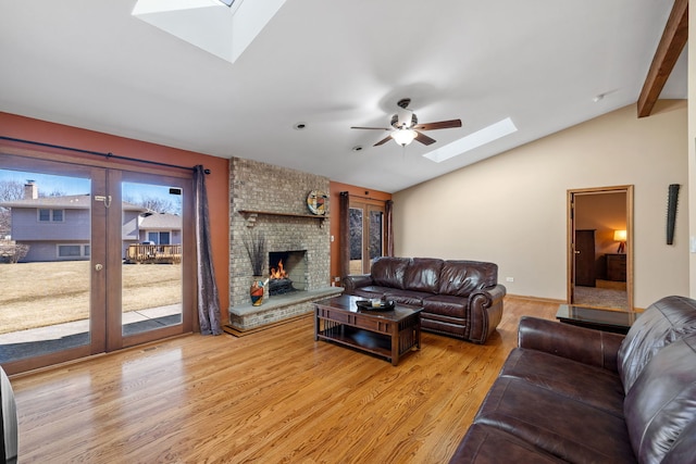 living room featuring vaulted ceiling with skylight, light wood-style flooring, a ceiling fan, french doors, and a brick fireplace