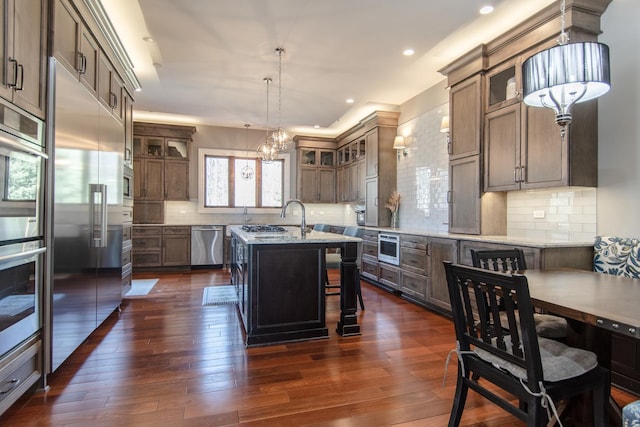 kitchen featuring a center island with sink, backsplash, dark wood finished floors, stainless steel appliances, and an inviting chandelier