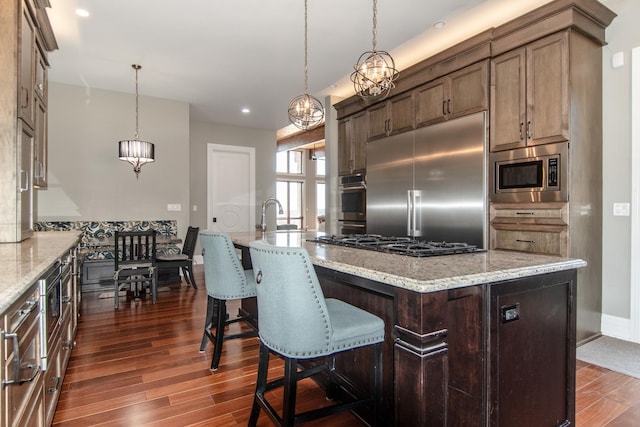 kitchen featuring dark wood-type flooring, an island with sink, a sink, built in appliances, and light stone countertops