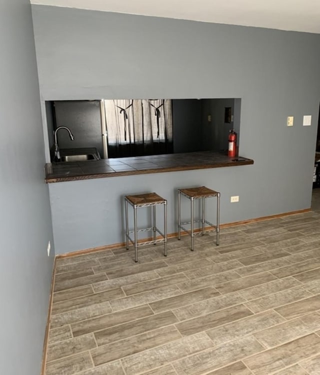 kitchen with tile counters, baseboards, a kitchen breakfast bar, wood tiled floor, and a sink