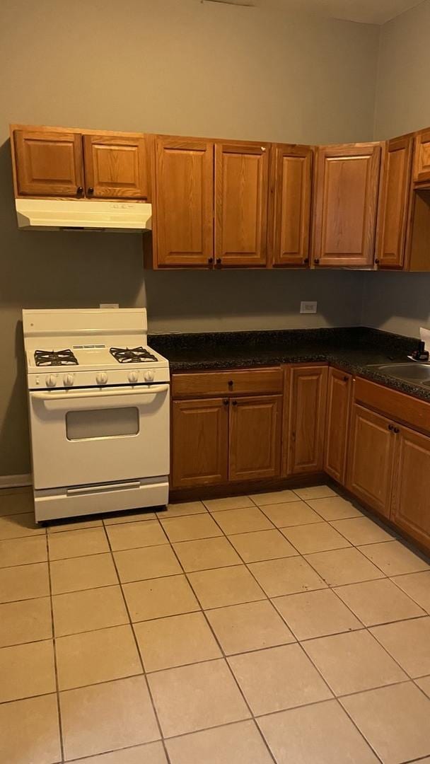 kitchen featuring light tile patterned flooring, under cabinet range hood, white range with gas stovetop, brown cabinetry, and dark countertops