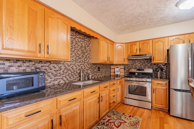 kitchen featuring under cabinet range hood, light wood-type flooring, dark stone countertops, appliances with stainless steel finishes, and a sink