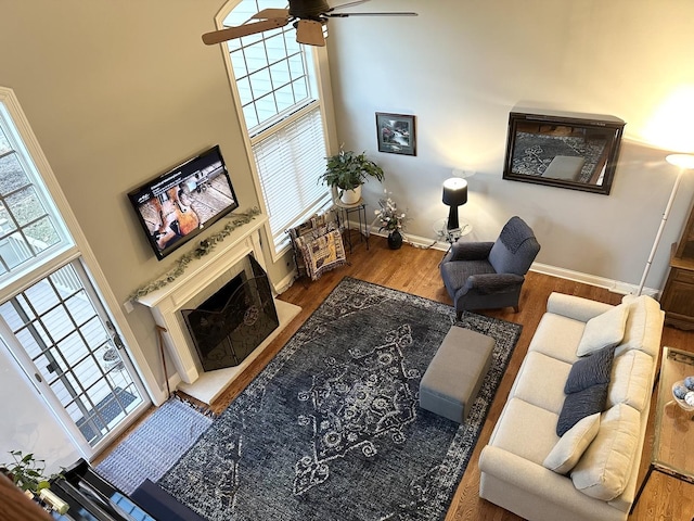 living room with ceiling fan, a fireplace with raised hearth, a high ceiling, wood finished floors, and baseboards