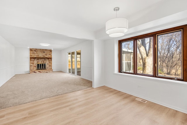 unfurnished living room with a wainscoted wall, visible vents, a brick fireplace, light colored carpet, and light wood-type flooring