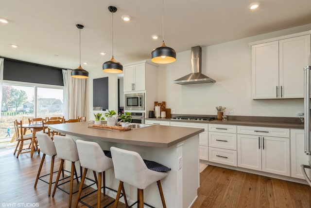kitchen with wood finished floors, a kitchen island, wall chimney exhaust hood, appliances with stainless steel finishes, and white cabinetry