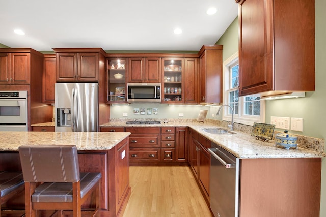 kitchen featuring light wood-style flooring, a sink, appliances with stainless steel finishes, glass insert cabinets, and light stone countertops