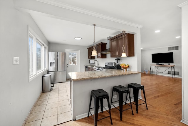 kitchen with appliances with stainless steel finishes, a peninsula, light stone countertops, dark brown cabinets, and a kitchen bar