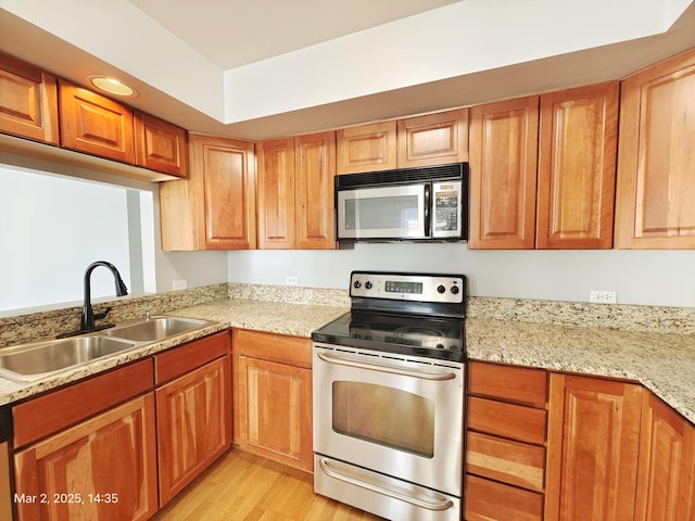 kitchen with brown cabinetry, light stone countertops, stainless steel appliances, light wood-style floors, and a sink