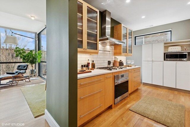 kitchen featuring wall chimney exhaust hood, light wood-style flooring, appliances with stainless steel finishes, light countertops, and backsplash