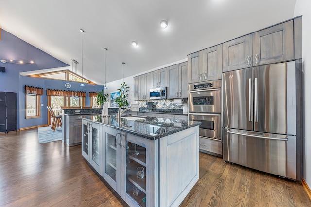 kitchen with dark wood-style floors, glass insert cabinets, appliances with stainless steel finishes, dark stone countertops, and vaulted ceiling