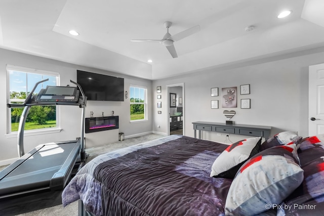 carpeted bedroom with a tray ceiling, multiple windows, a glass covered fireplace, and recessed lighting