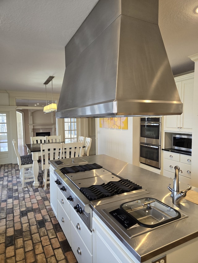 kitchen with brick floor, stainless steel appliances, a fireplace, white cabinets, and island exhaust hood