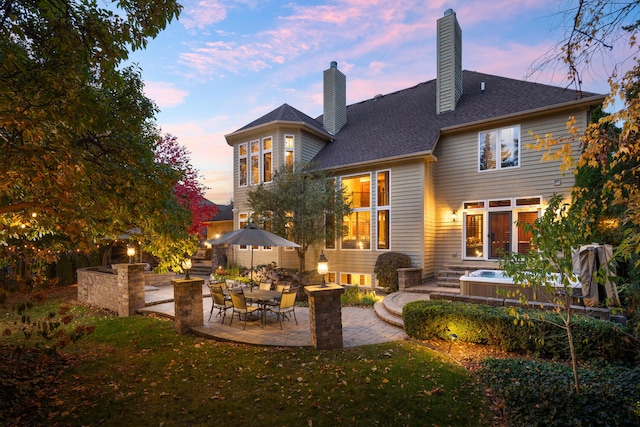 back of house at dusk with a yard, roof with shingles, a patio, and a chimney