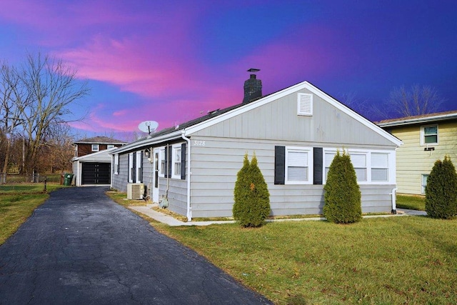 property exterior at dusk featuring driveway, a garage, a lawn, a chimney, and an outbuilding