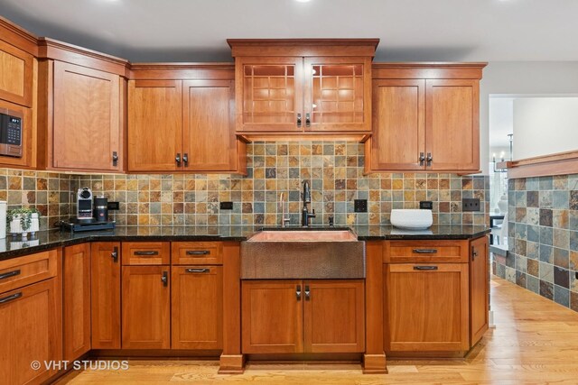 kitchen featuring light wood finished floors, decorative backsplash, brown cabinetry, dark stone countertops, and a sink