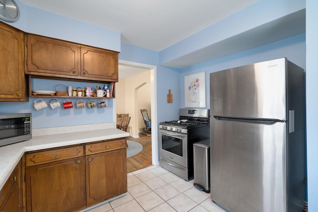 kitchen with light tile patterned floors, light countertops, appliances with stainless steel finishes, brown cabinets, and open shelves