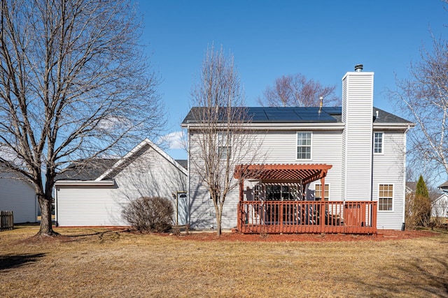 back of property with a yard, a wooden deck, roof mounted solar panels, a pergola, and a chimney