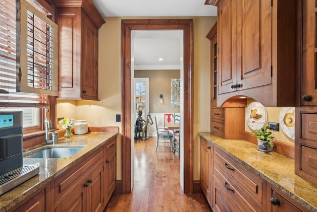 kitchen with brown cabinetry, ornamental molding, wood finished floors, light stone countertops, and a sink