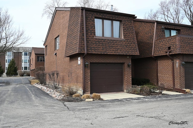 view of front of property featuring mansard roof, aphalt driveway, a garage, a shingled roof, and brick siding