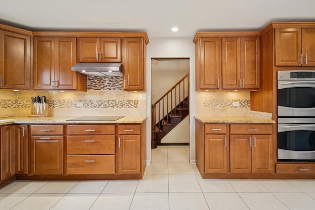 kitchen with under cabinet range hood, black electric stovetop, brown cabinetry, and stainless steel double oven