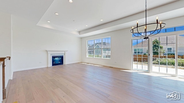 unfurnished living room with a notable chandelier, wood finished floors, baseboards, a tray ceiling, and a glass covered fireplace