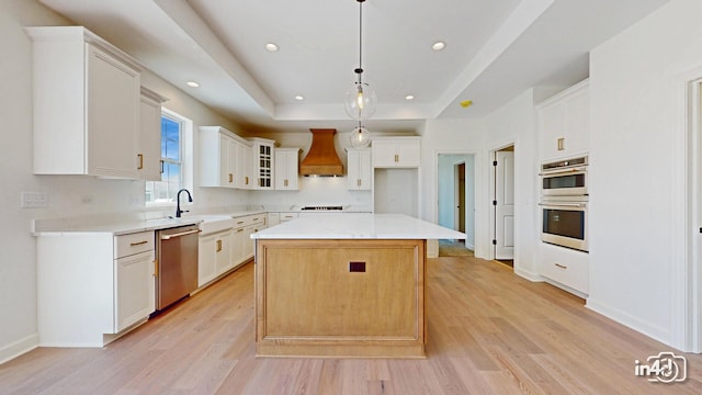 kitchen featuring light wood finished floors, a tray ceiling, appliances with stainless steel finishes, and custom range hood