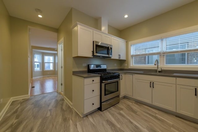 kitchen with dark countertops, appliances with stainless steel finishes, white cabinets, a sink, and light wood-type flooring