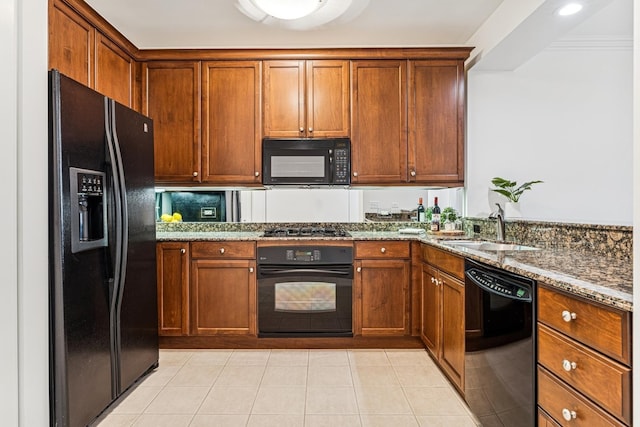 kitchen featuring light tile patterned floors, dark stone countertops, brown cabinetry, black appliances, and a sink