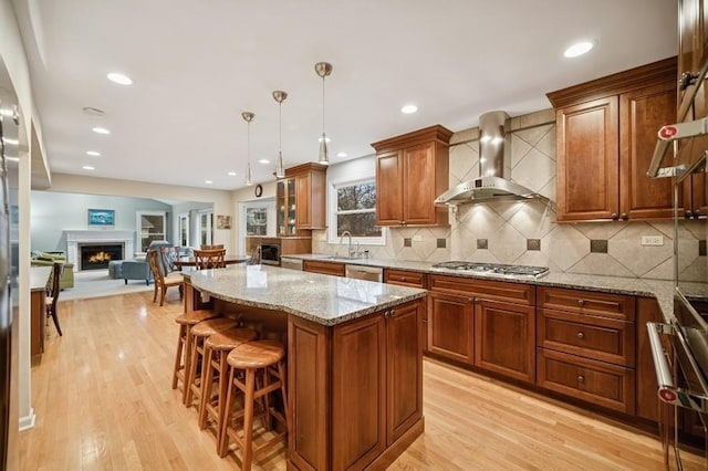 kitchen featuring a warm lit fireplace, wall chimney exhaust hood, appliances with stainless steel finishes, light wood-type flooring, and a sink