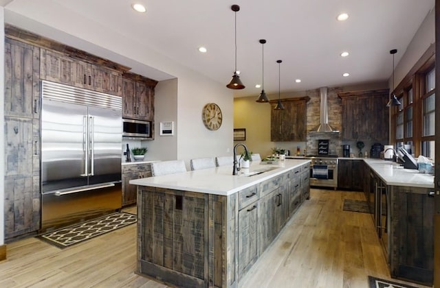 kitchen with a large island, a sink, built in appliances, light wood-type flooring, and wall chimney exhaust hood