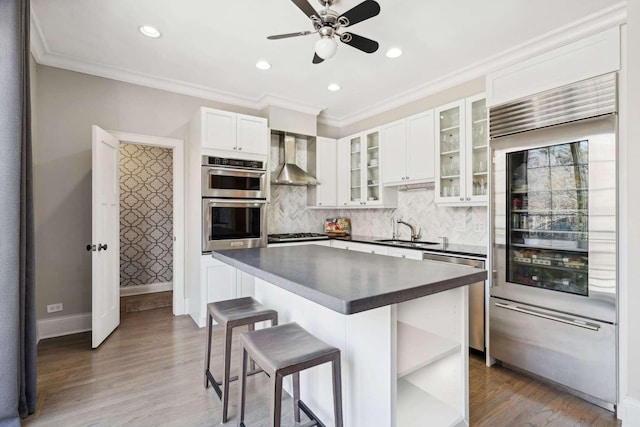 kitchen featuring wall chimney exhaust hood, open shelves, dark countertops, appliances with stainless steel finishes, and a kitchen bar