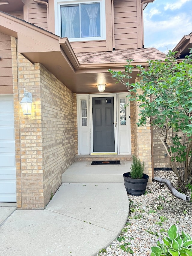 doorway to property with brick siding, an attached garage, and roof with shingles