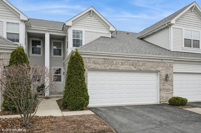 view of property with aphalt driveway, roof with shingles, brick siding, and a garage