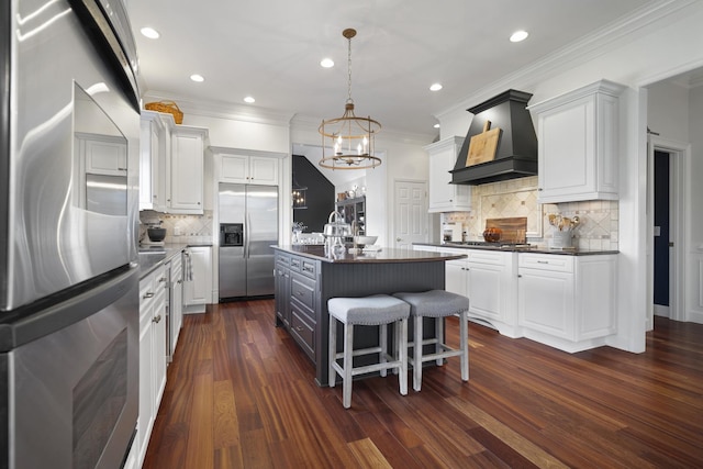 kitchen with stainless steel appliances, white cabinetry, dark countertops, a center island with sink, and custom range hood