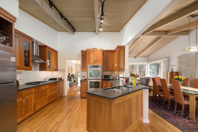 kitchen featuring brown cabinetry, dark countertops, wall chimney exhaust hood, stainless steel appliances, and a sink