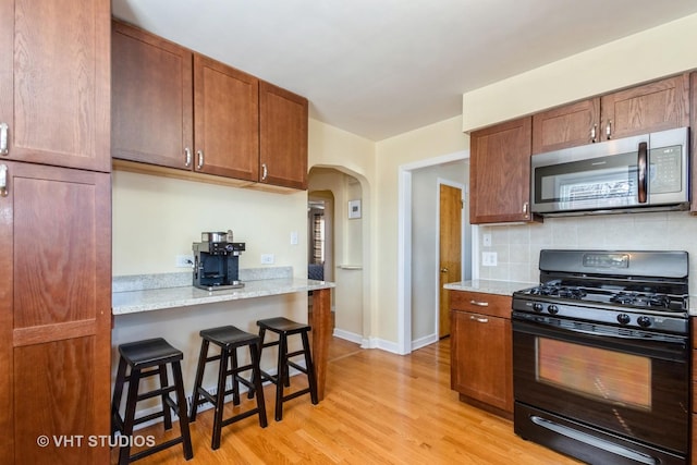 kitchen featuring light wood finished floors, stainless steel microwave, backsplash, black gas range, and a kitchen bar