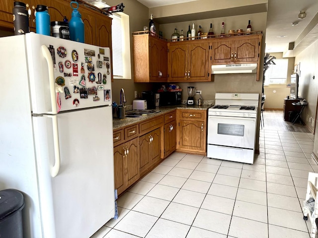 kitchen with light tile patterned floors, under cabinet range hood, white appliances, a sink, and brown cabinets