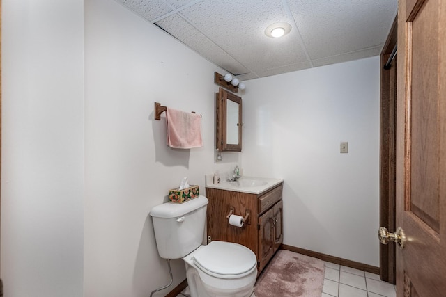 half bath with baseboards, a drop ceiling, toilet, tile patterned flooring, and vanity