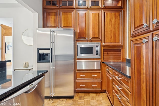 kitchen featuring stainless steel appliances, dark stone counters, brown cabinets, a warming drawer, and glass insert cabinets