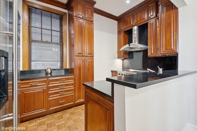 kitchen with a peninsula, wall chimney range hood, backsplash, brown cabinets, and dark countertops