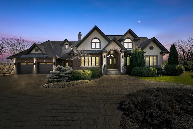 french country home featuring stone siding, a chimney, an attached garage, decorative driveway, and stucco siding