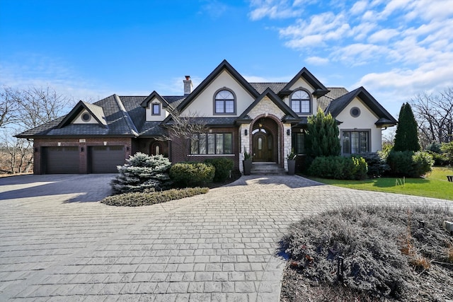 french country inspired facade featuring a garage, decorative driveway, stucco siding, a front lawn, and a chimney