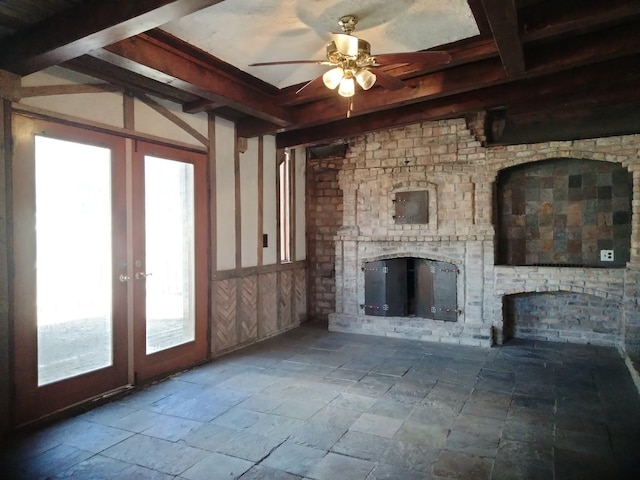 unfurnished living room with beamed ceiling, a ceiling fan, stone tile flooring, french doors, and a brick fireplace