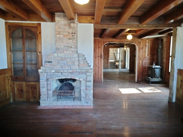 unfurnished living room featuring wooden walls, beamed ceiling, a wood stove, arched walkways, and wood-type flooring