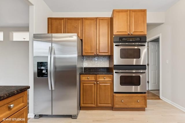 kitchen featuring decorative backsplash, brown cabinets, dark stone countertops, stainless steel appliances, and light wood-type flooring