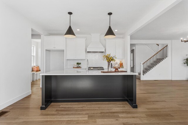 kitchen with light wood-style floors, custom exhaust hood, a center island with sink, and light countertops