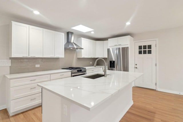 kitchen with wall chimney range hood, light wood-style flooring, appliances with stainless steel finishes, white cabinets, and a sink