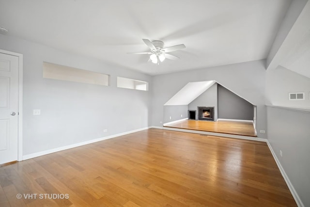 additional living space with visible vents, a ceiling fan, wood finished floors, a warm lit fireplace, and baseboards